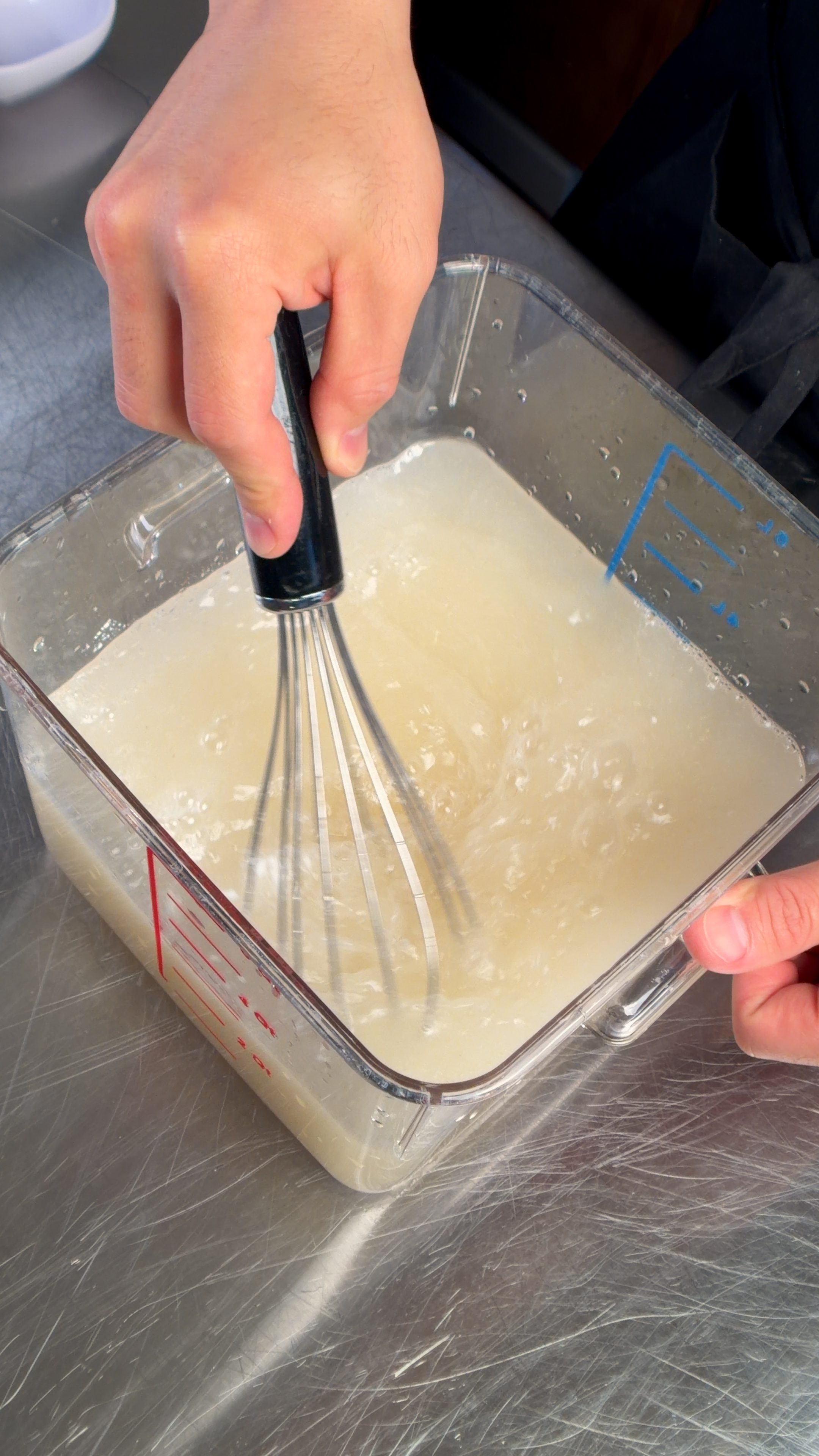 Brine being whisked in a clear square container on a stainless steel surface