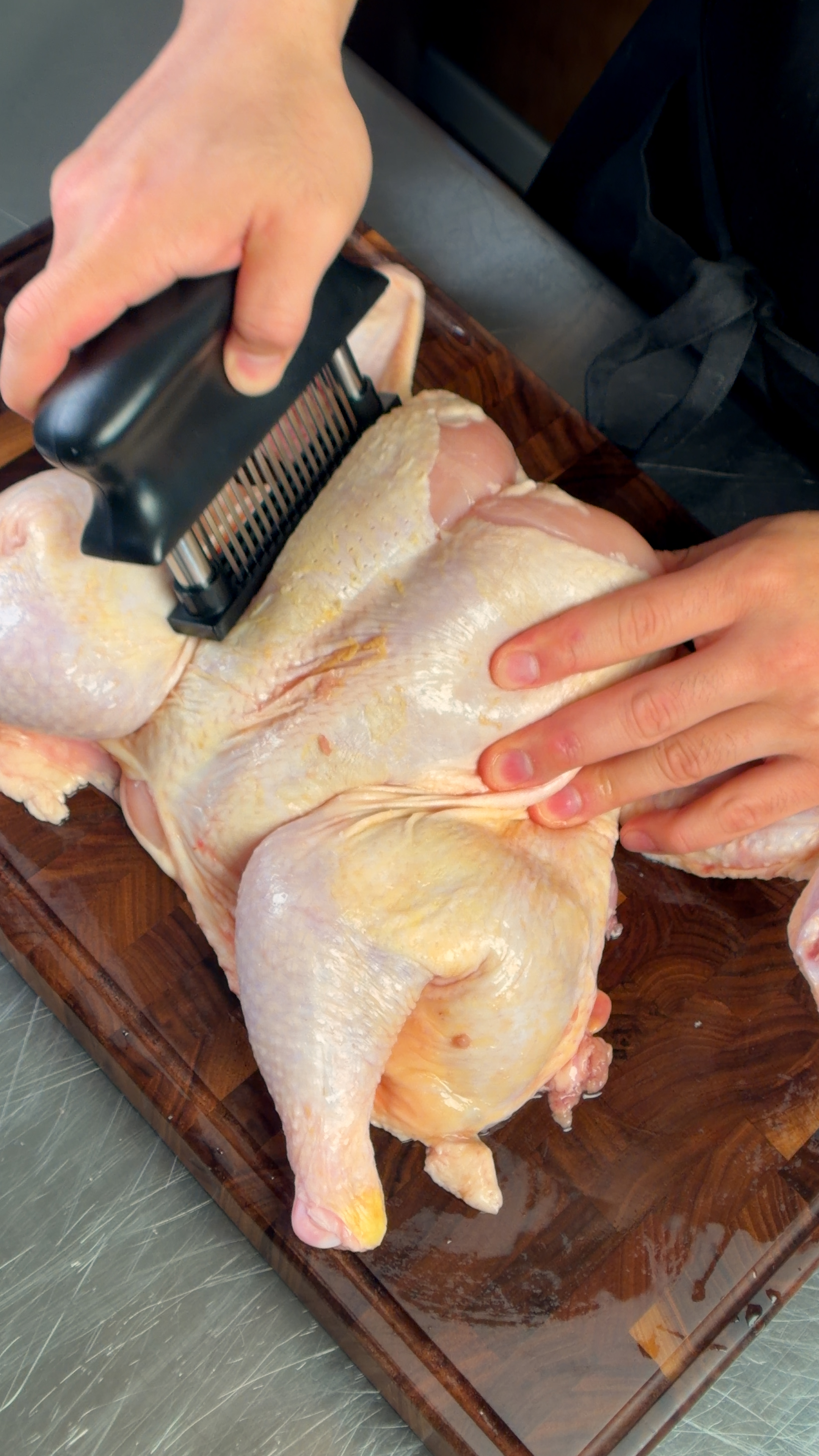 Hands using a needle meat tenderizer on a spatchcocked raw chicken on a wooden cutting board