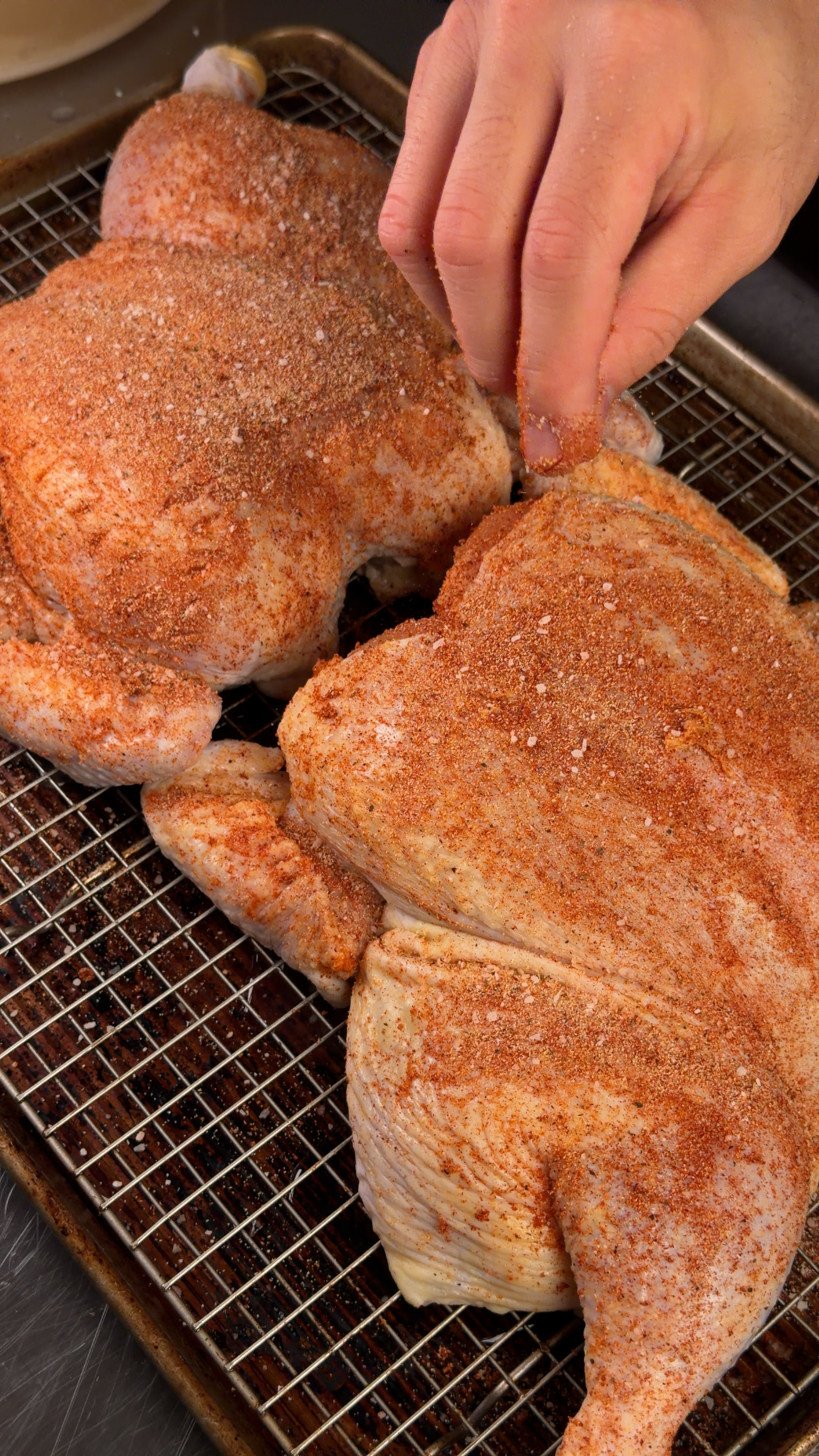 Two spatchcocked chickens generously coated in red spice rub resting uncovered on a wire rack over a baking sheet