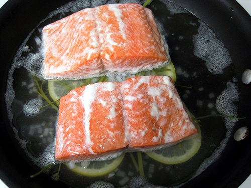 Two salmon fillets cooking in liquid with white albumin foaming out across the surface
