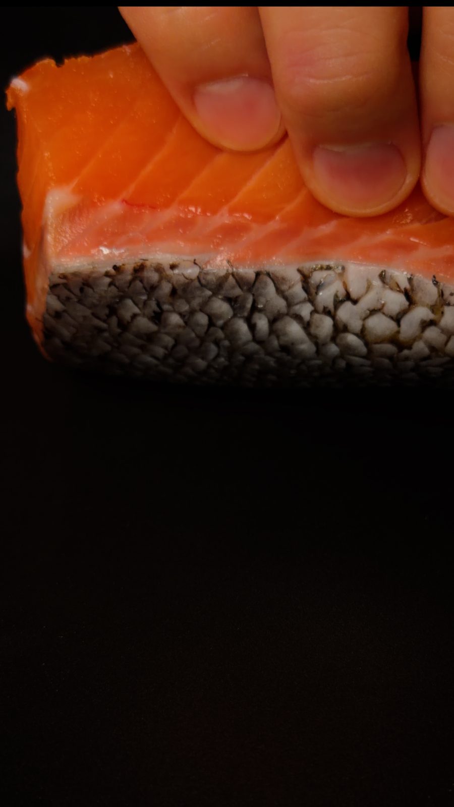 A close-up of the cut edge of a salmon fillet showing the orange flesh, a thin pale fat layer, and the skin