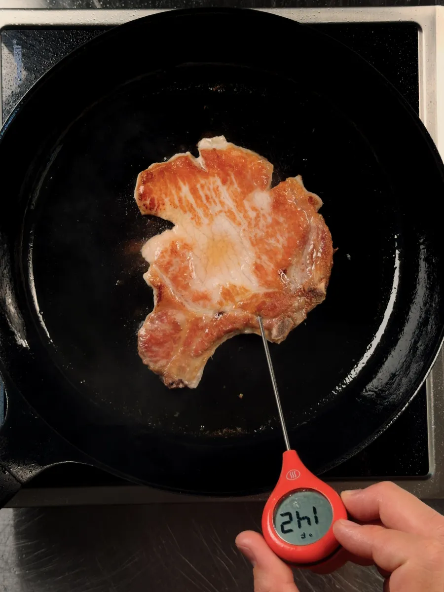A seared pork chop in a cast iron skillet with an instant-read thermometer reading 142°F