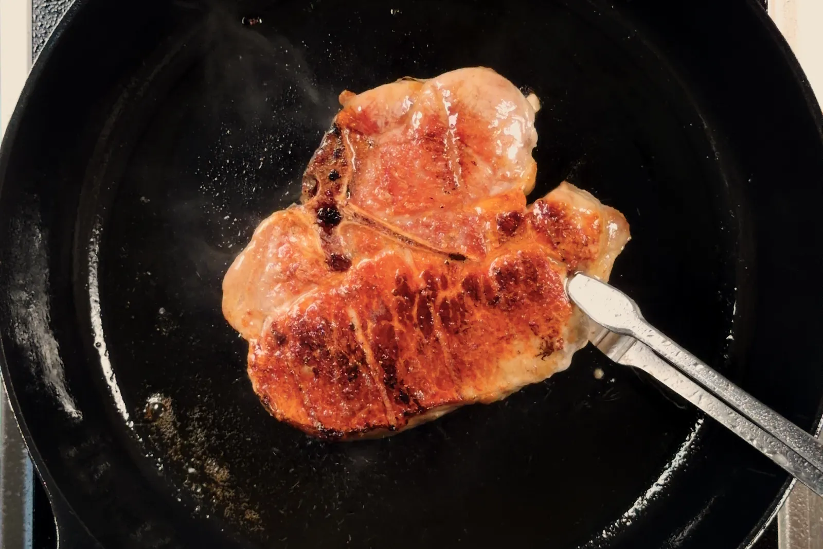 A deeply seared pork chop resting in a cast iron skillet, tongs alongside