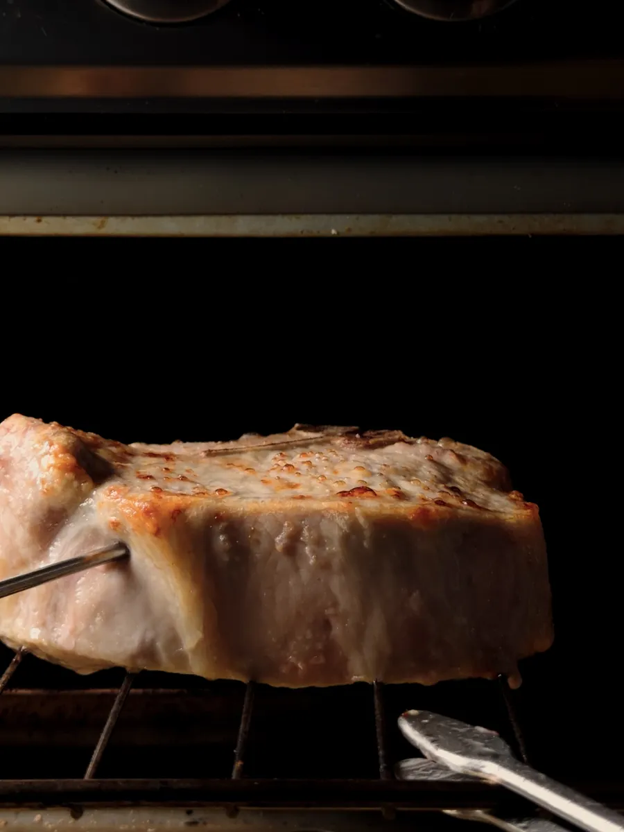 A thick pork chop on an oven rack with a probe thermometer inserted, oven dials above