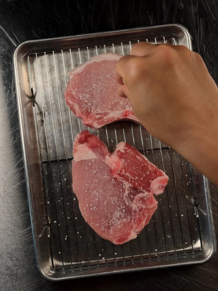 A thick bone-in pork chop and a thinner chop side by side on a wire rack, being salted