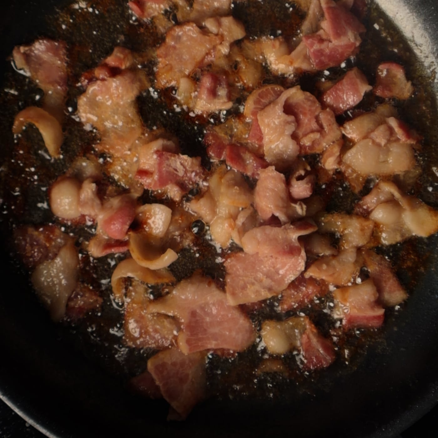 Bacon pieces rendering in a hot pan, releasing pools of golden fat