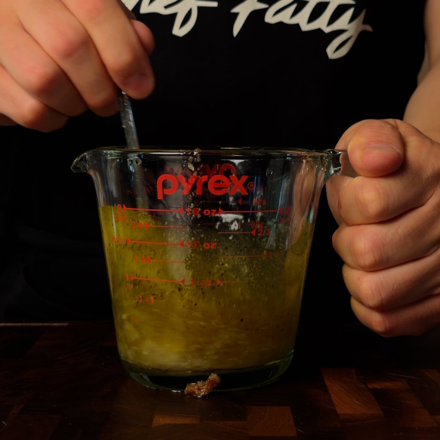 Pyrex measuring cup with golden roasted garlic vinaigrette and cracked black pepper visible, Chef Fatty apron in background