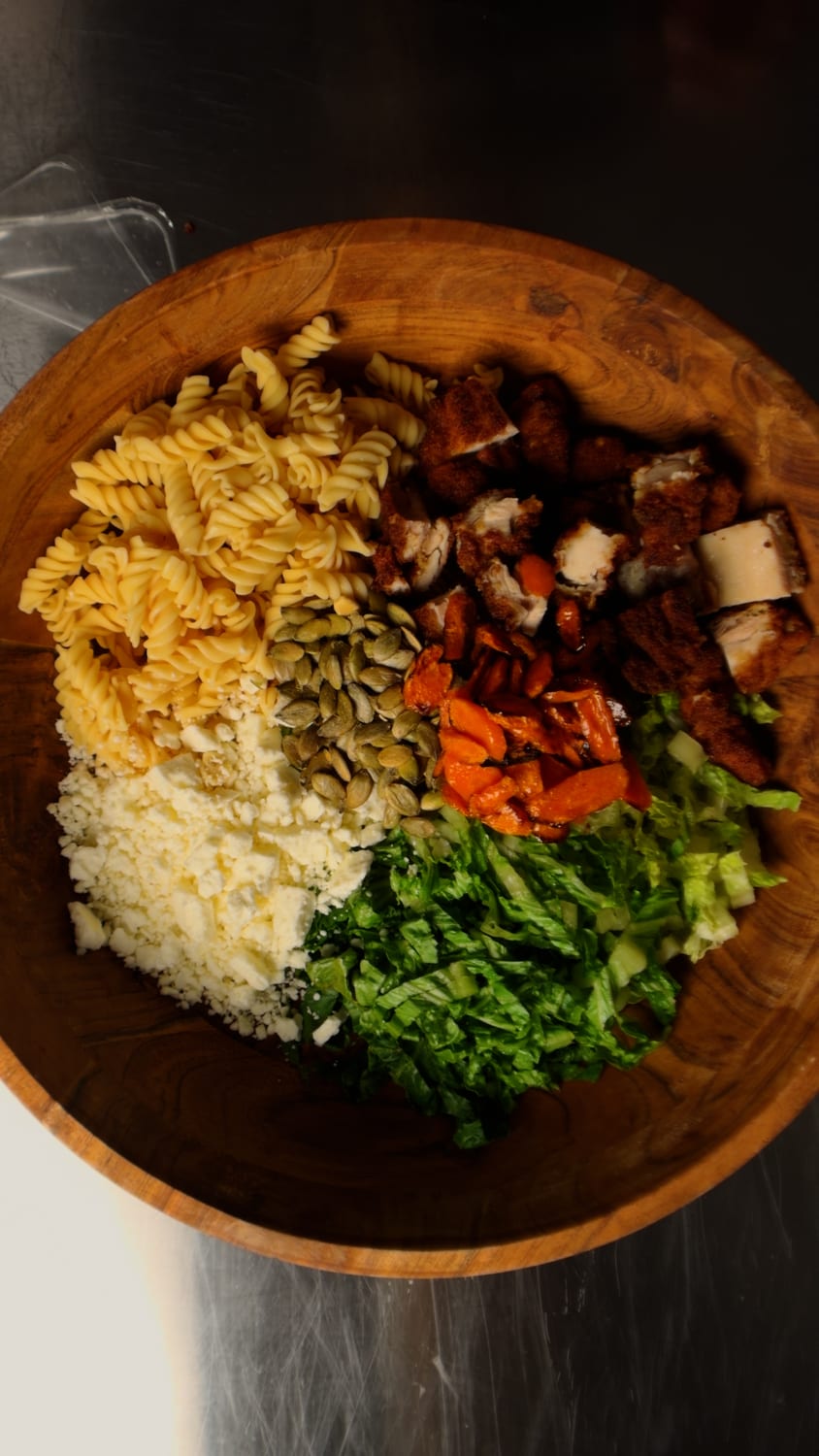 A wooden salad bowl with sections of rotini, crispy breaded chicken, roasted carrot coins, crumbled feta, toasted pepitas, and chopped greens pre-toss