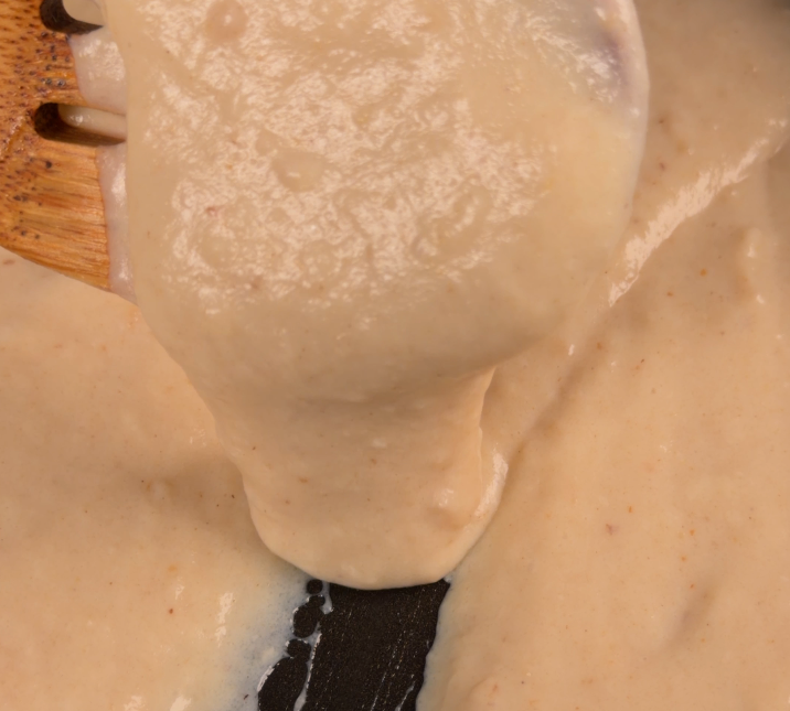A wooden spoon lifting smooth white sauce that ribbons off in a thick glossy stream